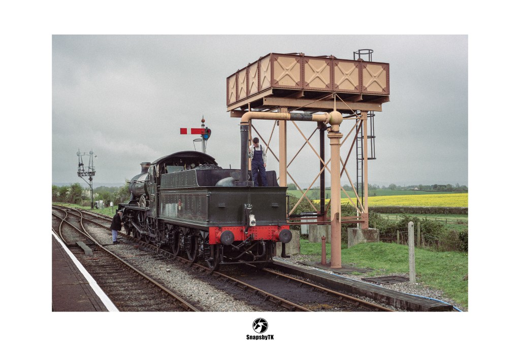 A vintage steam locomotive being refilled at an elevated water tower beside a yellow field.