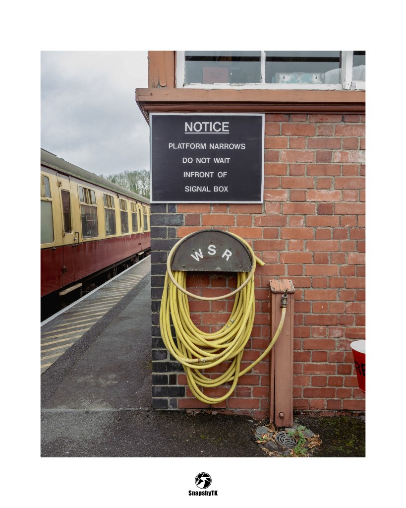 Sign reading 'NOTICE PLATFORM NARROWS DO NOT WAIT INFRONT OF SIGNAL BOX' on a brick wall beside a train.